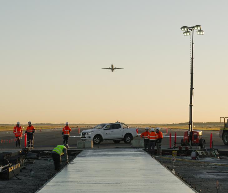 Construction workers pouring concrete on the Legacy Runway with a plane flying overhead